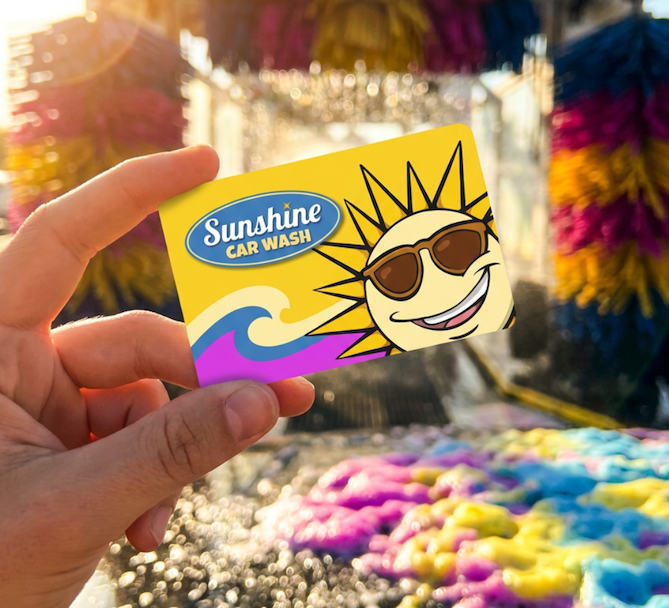 A hand holding a Sunshine Car Wash gift card in front of the colorful wash tunnel