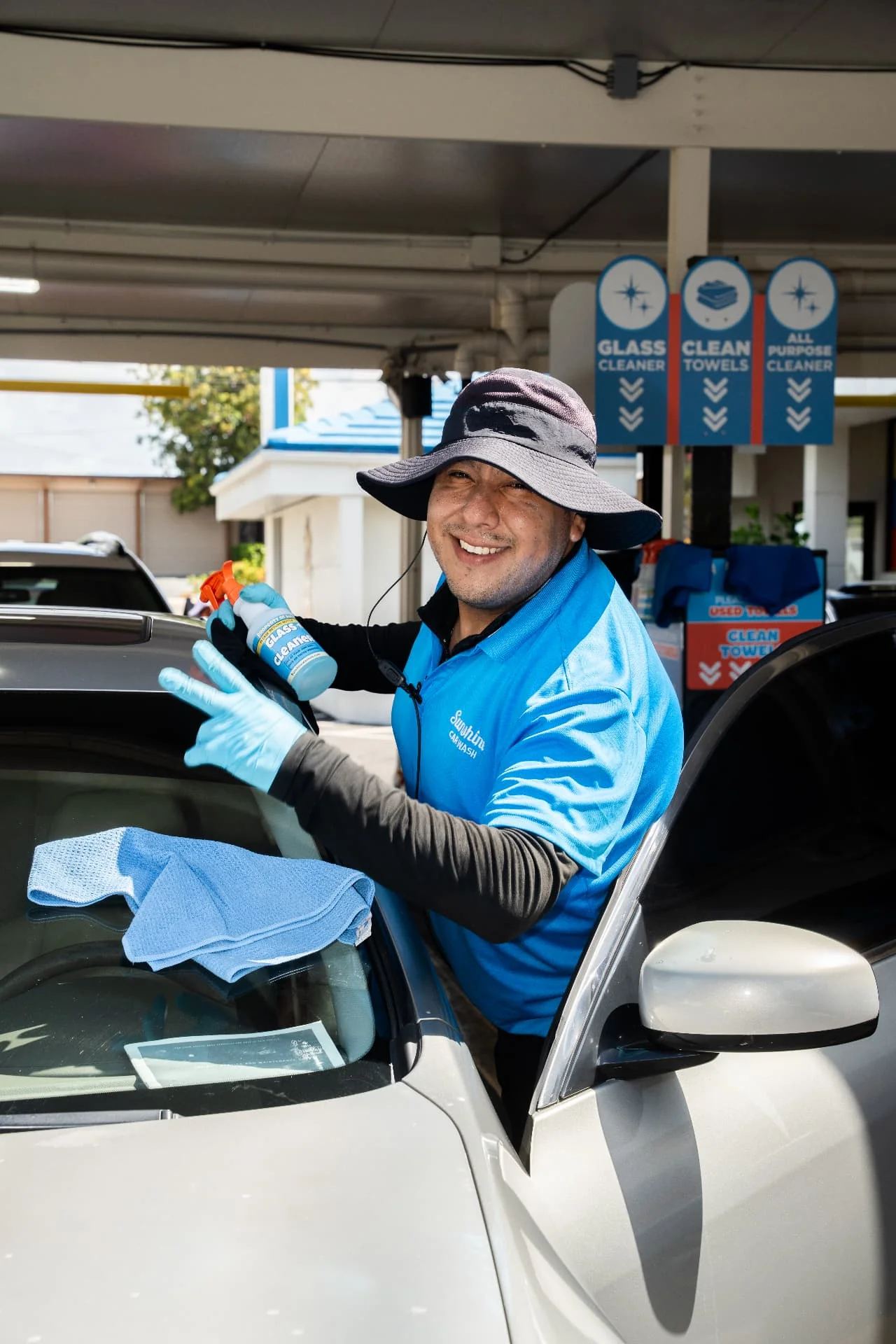 A smiling Sunshine team member in a blue uniform cleaning a car windshield with glass cleaner