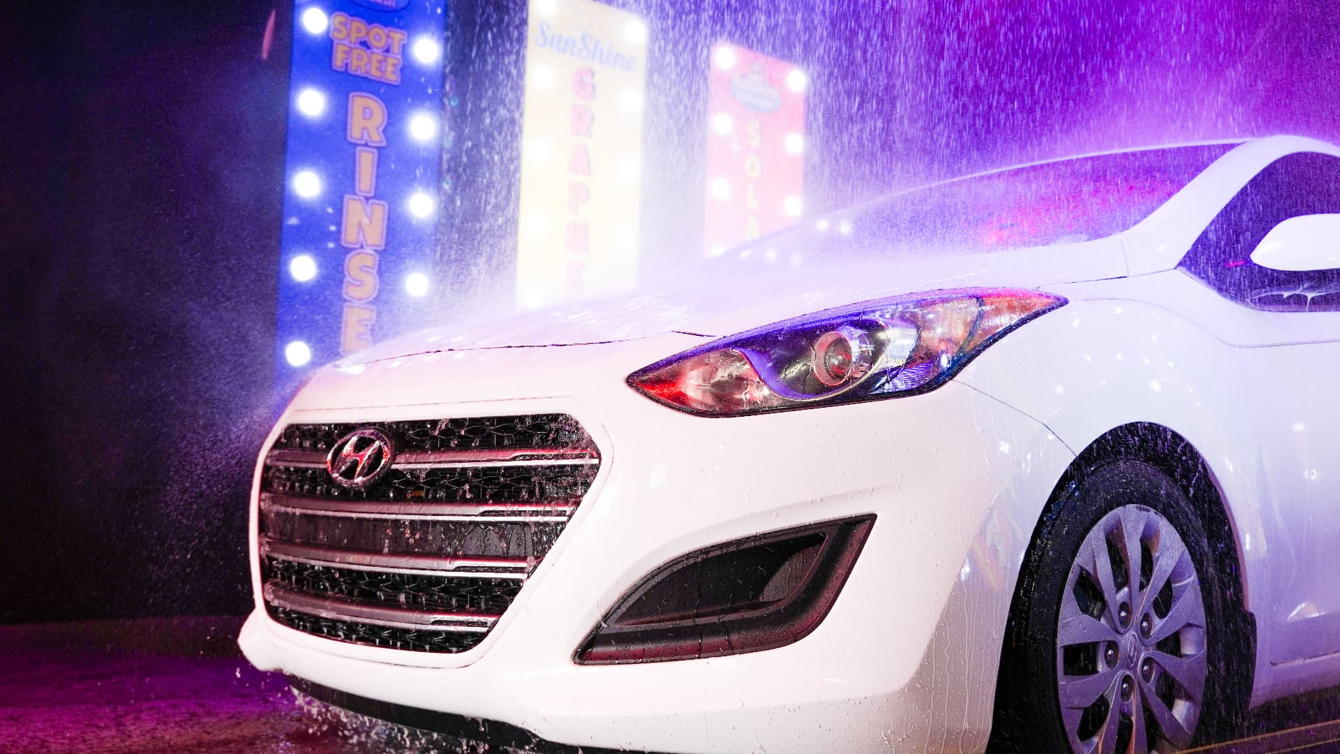A white car rolling through the glowing Sunshine Car Wash tunnel under purple and pink lights